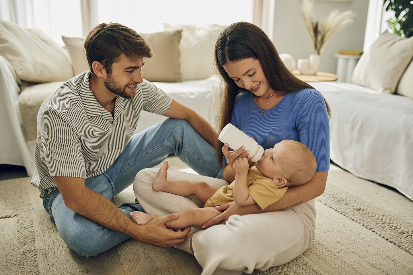 Beba pije mlijeko iz bočice dok je majka drži u naručju, a otac sjedi pored nje