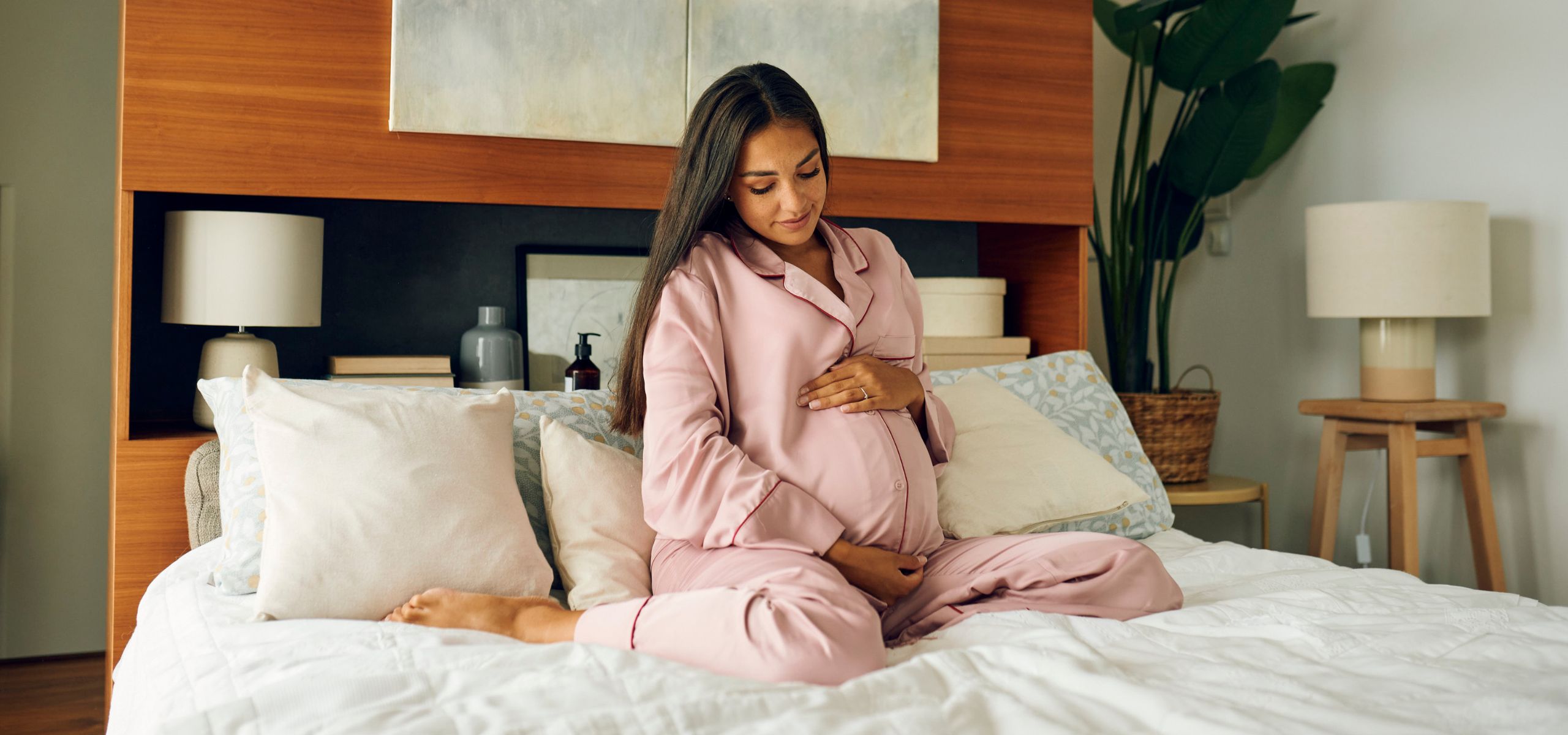 Trudnica u ružičastom ogrtaču sjedi na krevetu i drži se za trbuh.